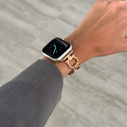 Rose gold circle link Apple Watch band worn on a wrist above grey trousers. Slim metal iWatch strap for everyday wear, compatible with Apple Watch 38mm to 49mm, Series 1-11, SE and Ultra