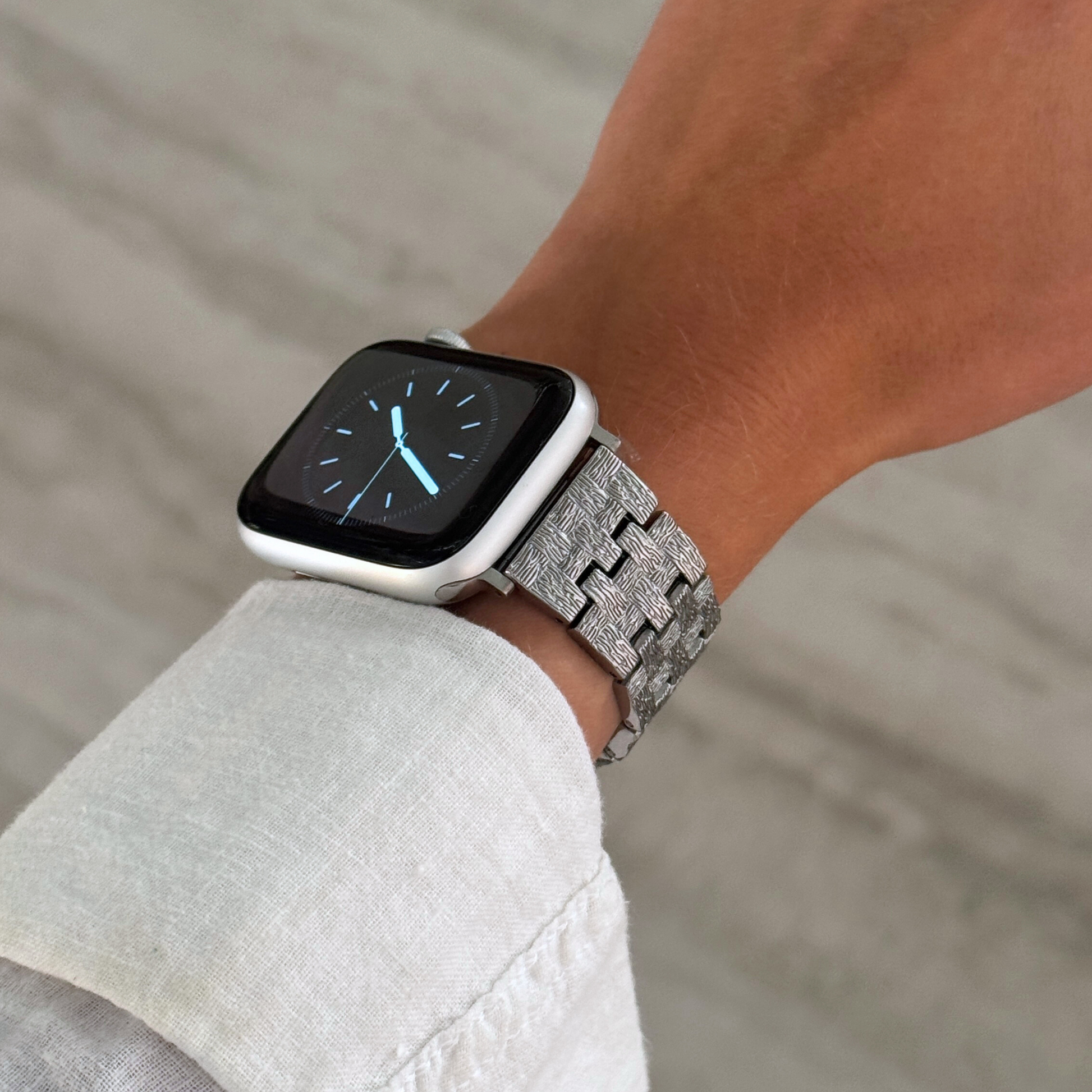 Silver baroque pattern Apple Watch band worn on a wrist with a pale linen sleeve. Luxurious metal iWatch strap with an intricate pattern, compatible with Apple Watch 38mm to 49mm, Series 1-11, SE and Ultra