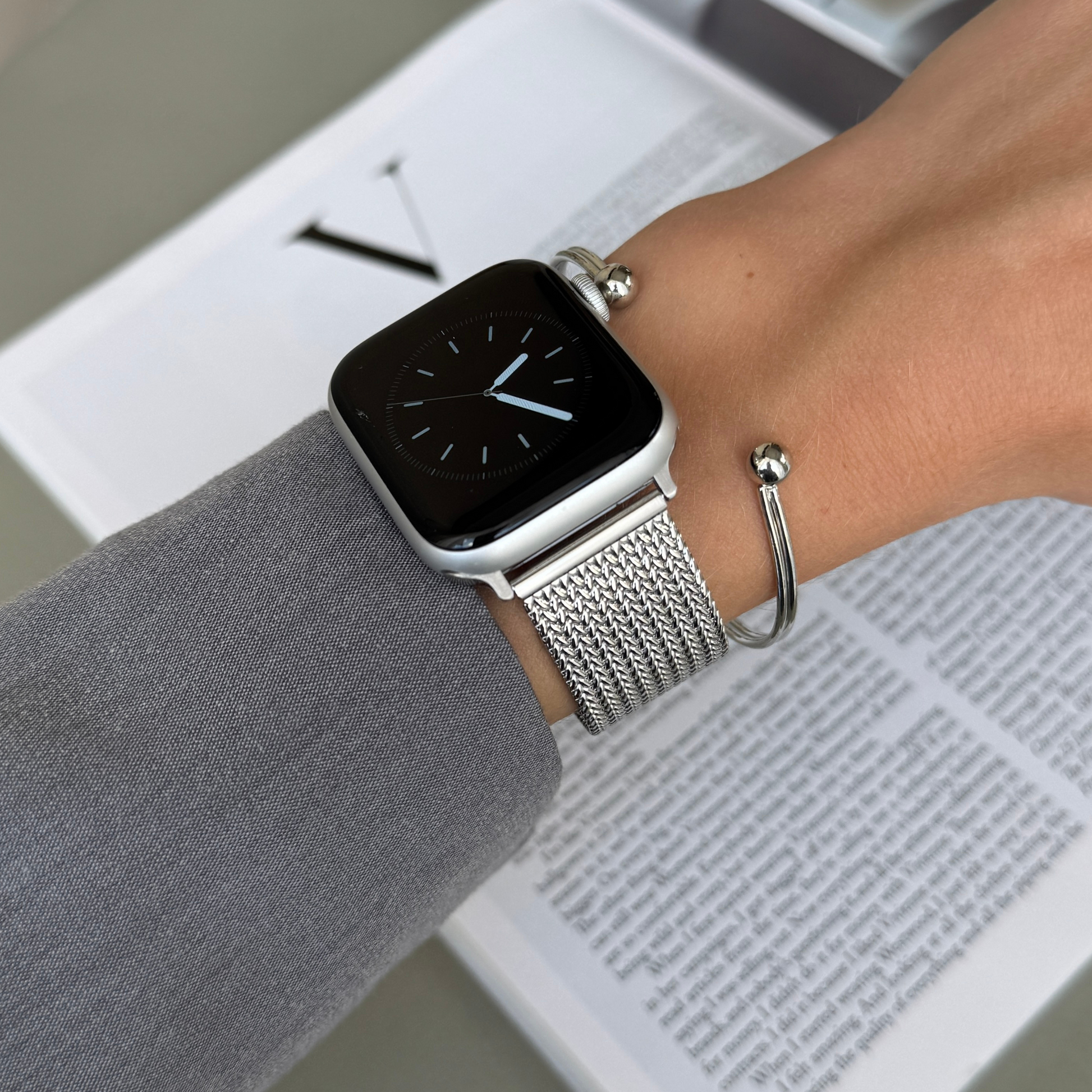 Silver mesh loop Apple Watch band worn with a silver bangle beside a magazine. Unisex stainless steel iWatch strap, compatible with Apple Watch 38mm to 49mm, Series 1-11, SE and Ultra