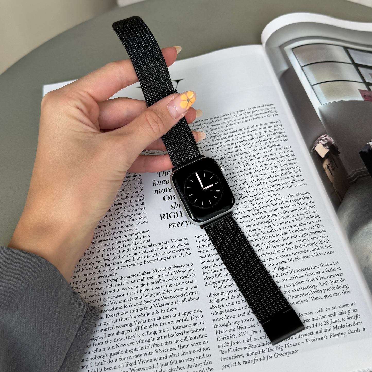 Black mesh loop Apple Watch band held in a hand over a magazine page. Everyday stainless steel iWatch bracelet, compatible with Apple Watch 38mm to 49mm, Series 1-11, SE and Ultra