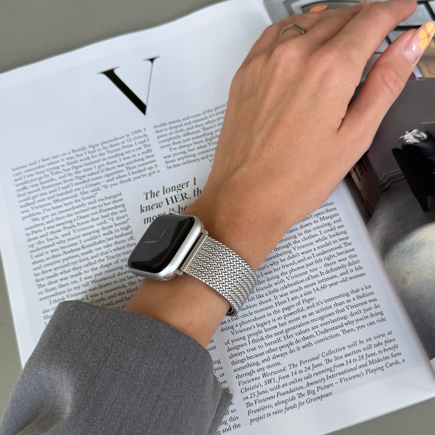 Starlight mesh loop Apple Watch band worn on a wrist reaching across a magazine. Minimalist stainless steel iWatch strap, compatible with Apple Watch 38mm to 49mm, Series 1-11, SE and Ultra