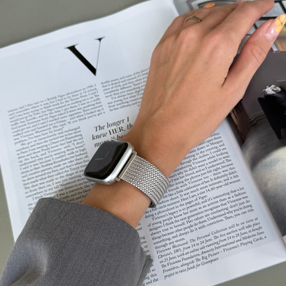 Starlight mesh loop Apple Watch band worn on a wrist reaching across a magazine. Minimalist stainless steel iWatch strap, compatible with Apple Watch 38mm to 49mm, Series 1-11, SE and Ultra