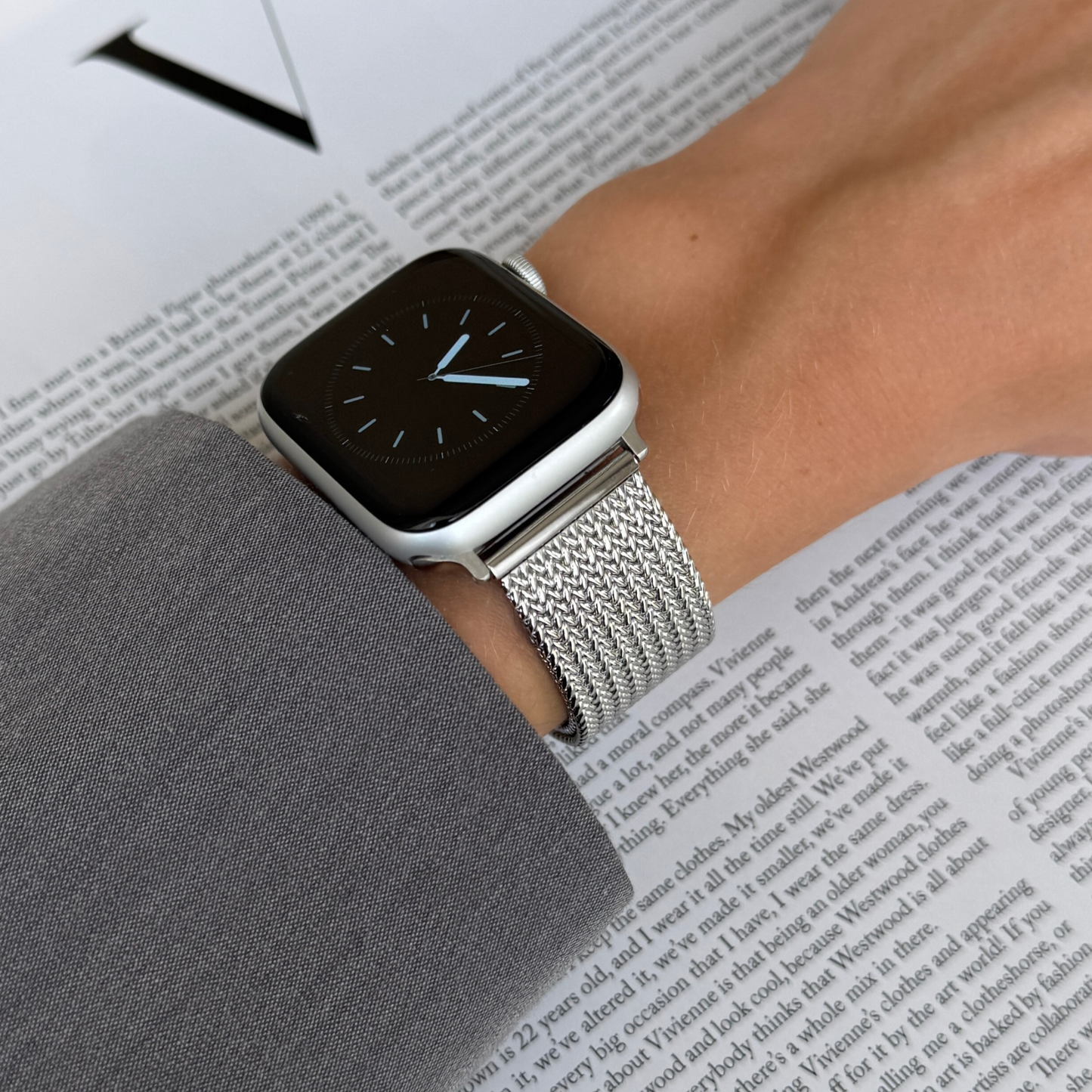 Silver mesh loop Apple Watch band worn on a wrist above an open magazine. Waterproof stainless steel iWatch bracelet, compatible with Apple Watch 38mm to 49mm, Series 1-11, SE and Ultra
