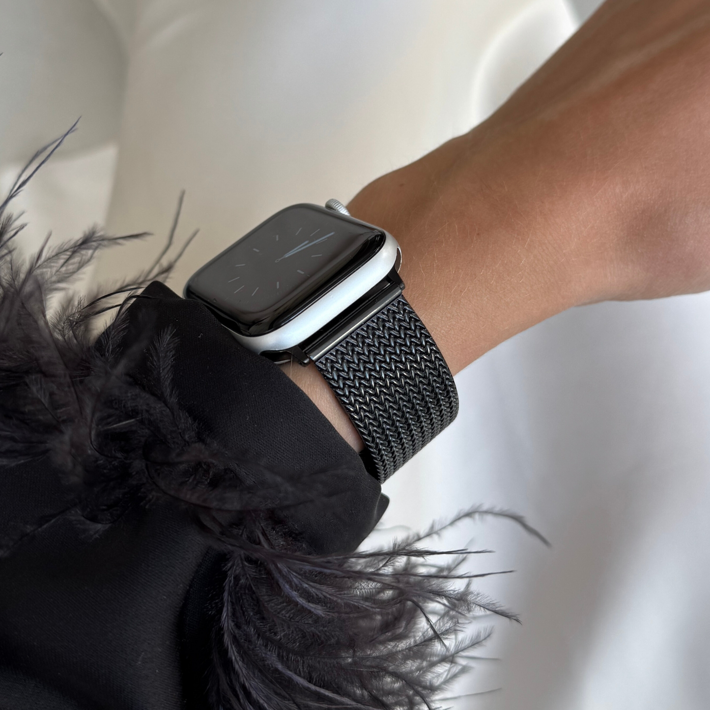 Black mesh loop Apple Watch band worn with a feather trimmed cuff above white satin. Unisex stainless steel iWatch strap, compatible with Apple Watch 38mm to 49mm, Series 1-11, SE and Ultra