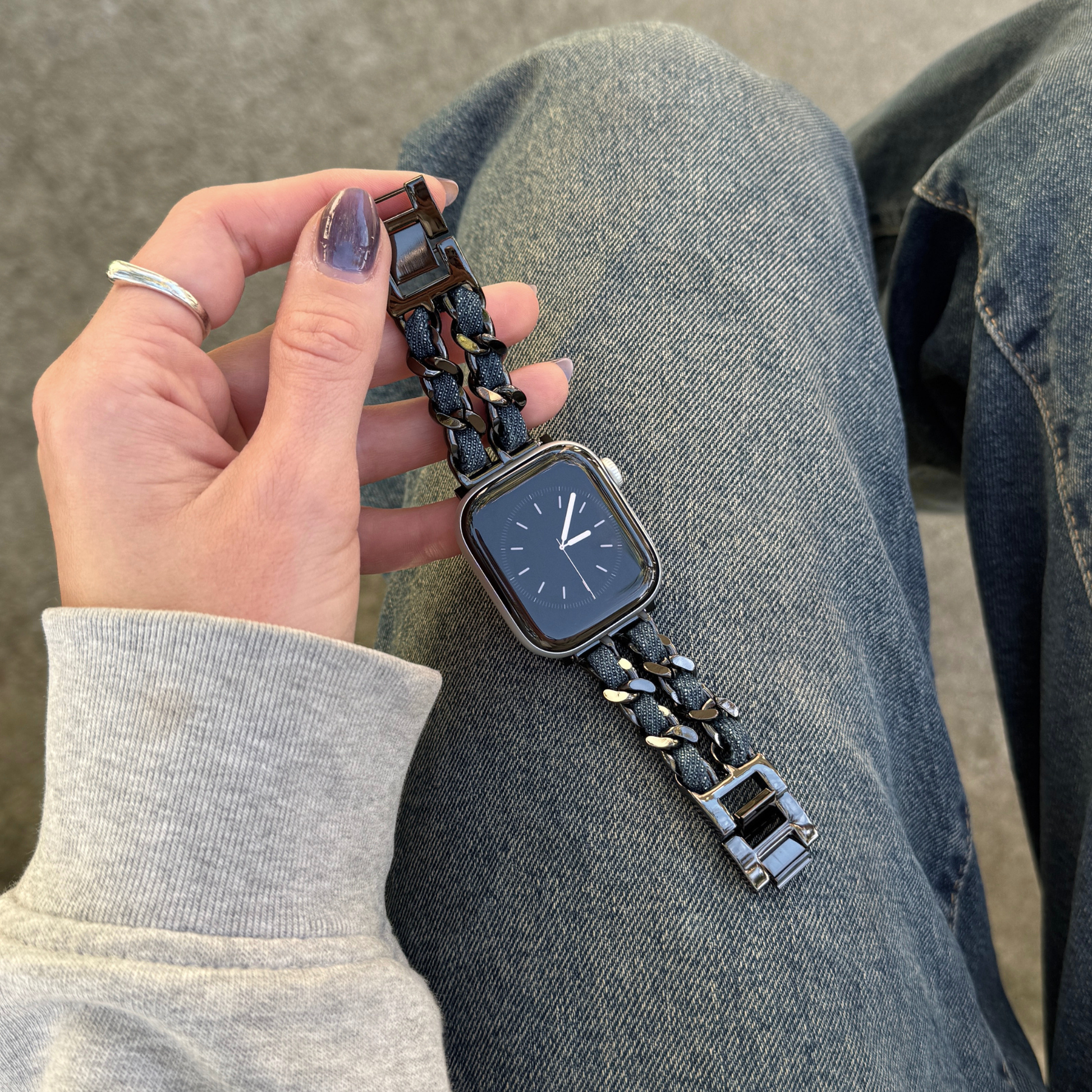 A close-up of a person's hand holding a an Apple Watch with a black metallic link band, set against their denim-clad legs and concrete floor.