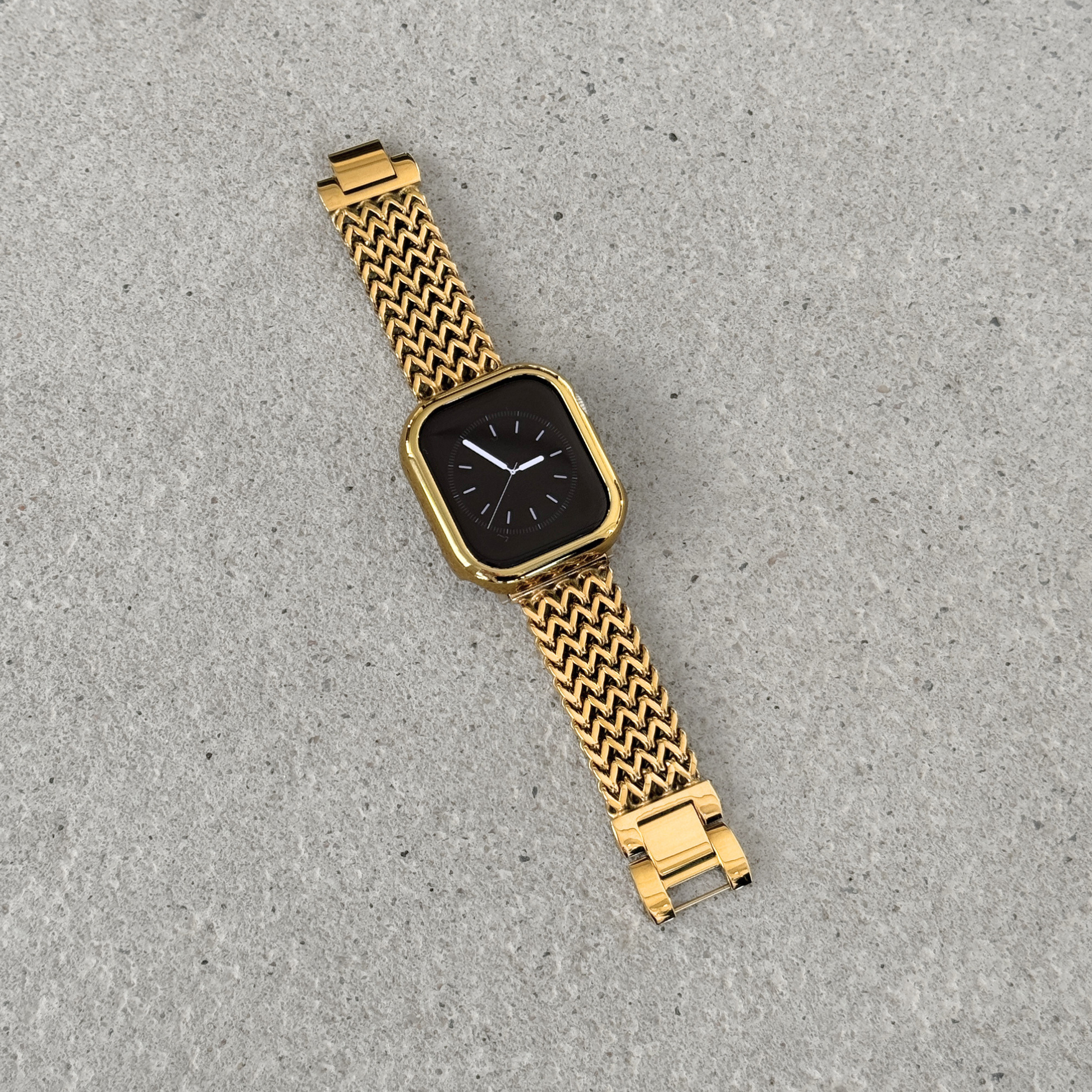 Apple Watch with a gold bumper case cover and a woven metallic mesh strap, photographed diagonally on a textured surface.