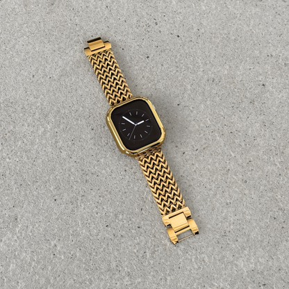 Apple Watch with a gold bumper case cover and a woven metallic mesh strap, photographed diagonally on a textured surface.