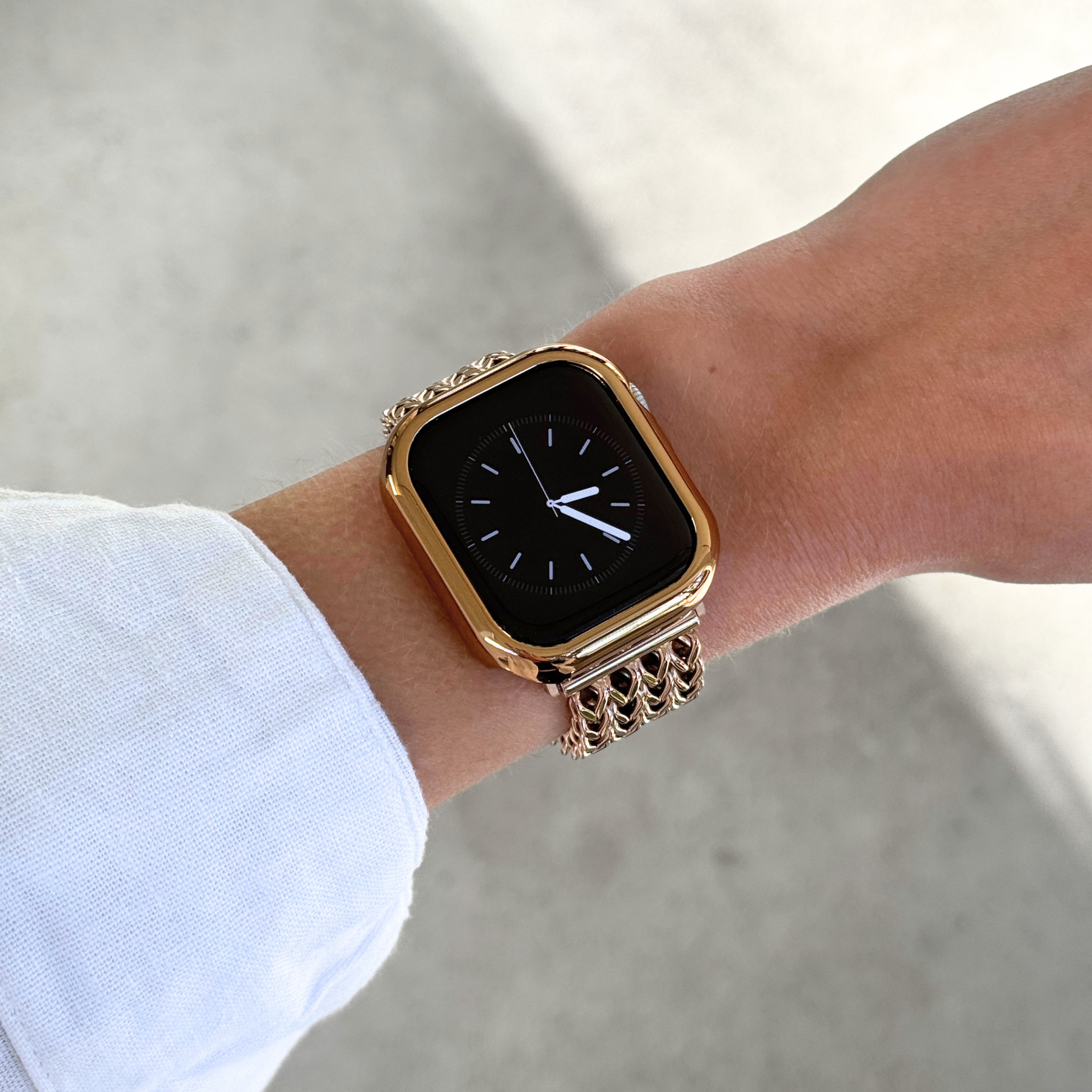 A close-up of an Apple Watch with a rose gold toned bezel, bumper case cover, with a matching chain-link band worn on a person's wrist, set against a soft concrete background.