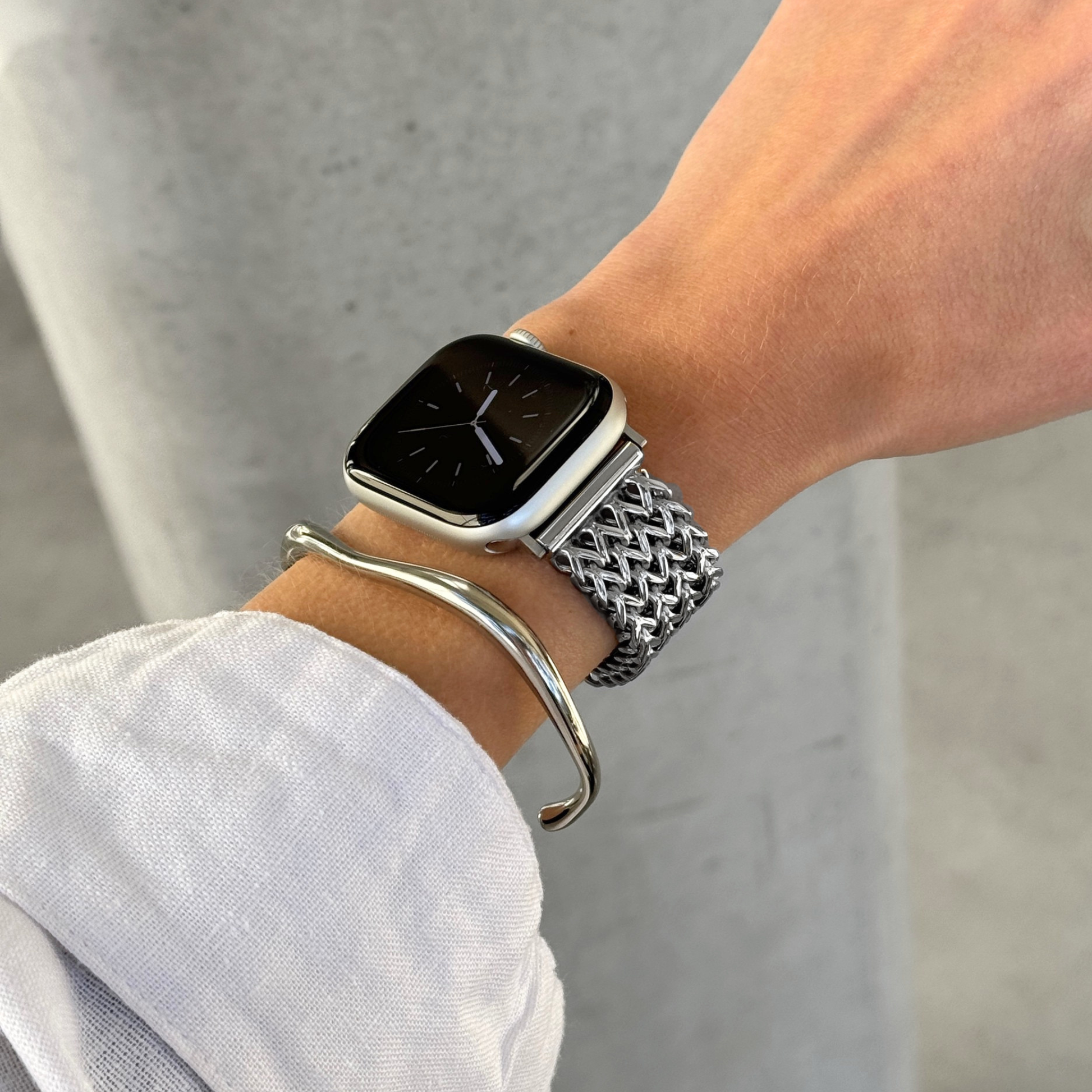 A close-up of a person's wrist wearing an Apple smartwatch with a silver mesh band and a silver curved bangle bracelet, against a light background.