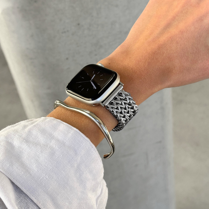 A close-up of a person's wrist wearing an Apple smartwatch with a silver mesh band and a silver curved bangle bracelet, against a light background.