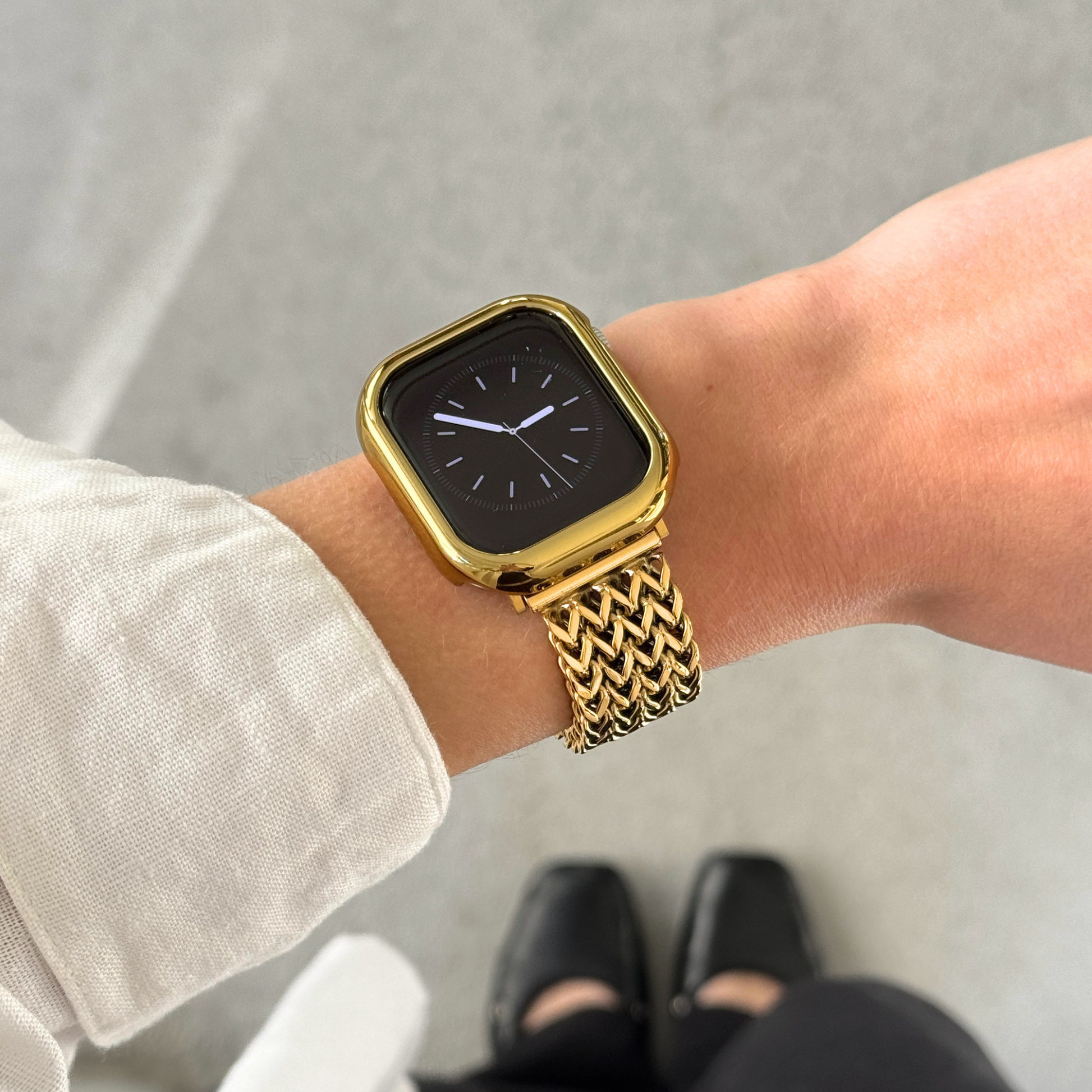 Apple watch with a gold bumper case and a woven metallic gold band, shown worn on a person's wrist against a light concrete floor background.