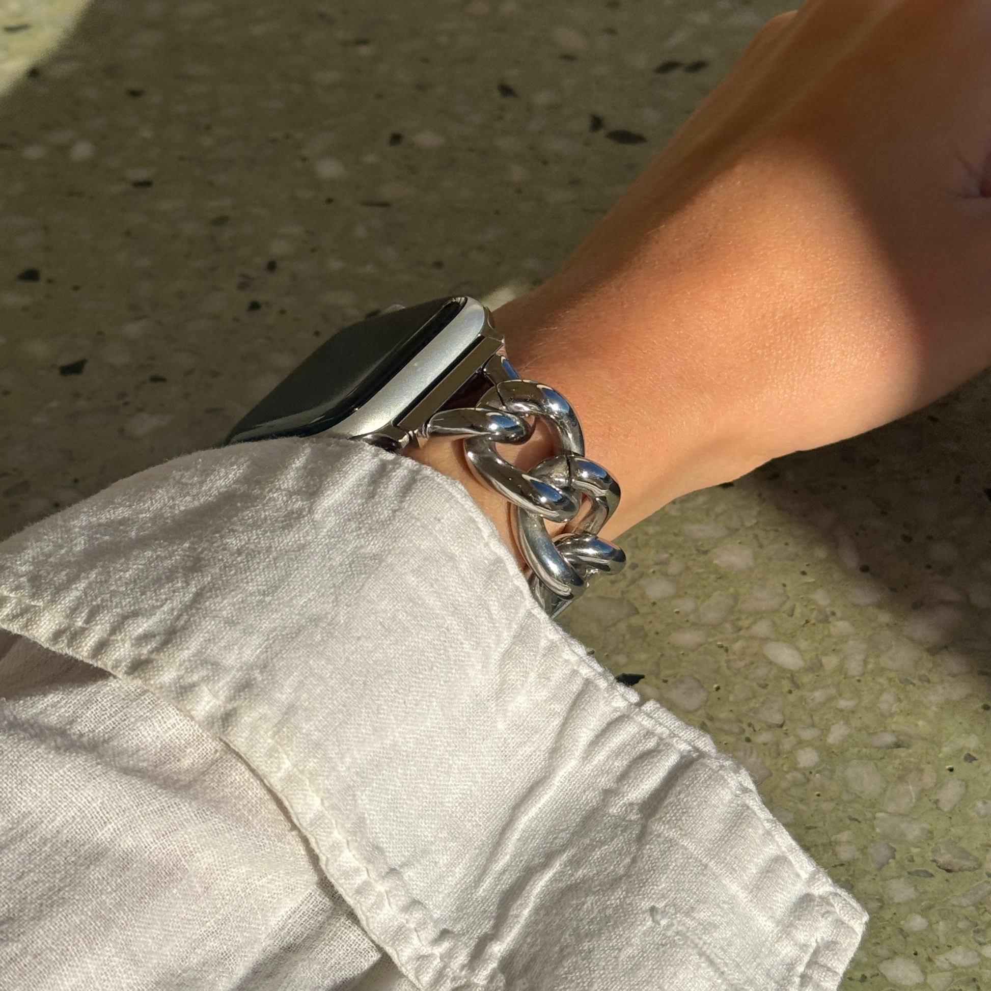 Person wearing a smartwatch with a chain bracelet Silver chain link Apple Watch band worn on a wrist with a cream linen sleeve. Fashion forward stainless steel iWatch bracelet, compatible with Apple Watch 38mm to 49mm, Series 1-11, SE and Ultrat on a concrete surface