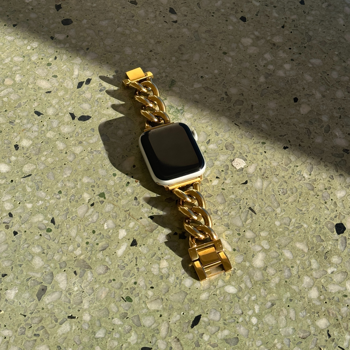 Gold chain link Apple Watch band lying in sunlight on a terrazzo surface. Beautiful stainless steel iWatch bracelet, compatible with Apple Watch 38mm to 49mm, Series 1-11, SE and Ultra