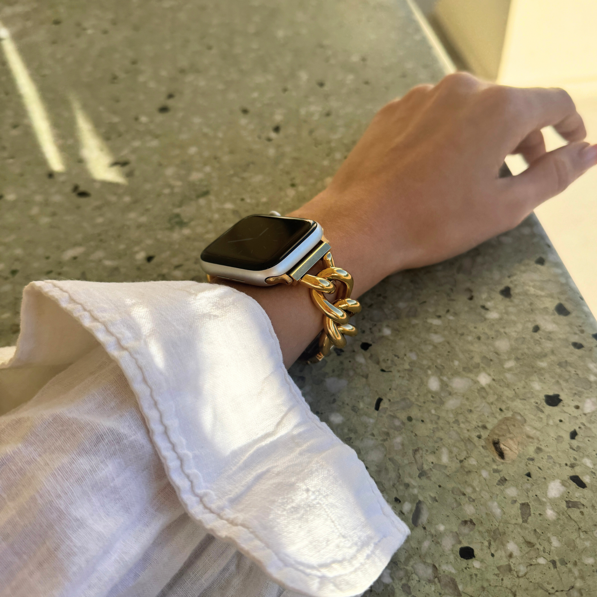 Gold chain link Apple Watch band worn with a white blouse in bright sunlight. Fashion forward stainless steel iWatch strap, compatible with Apple Watch 38mm to 49mm, Series 1-11, SE and Ultra