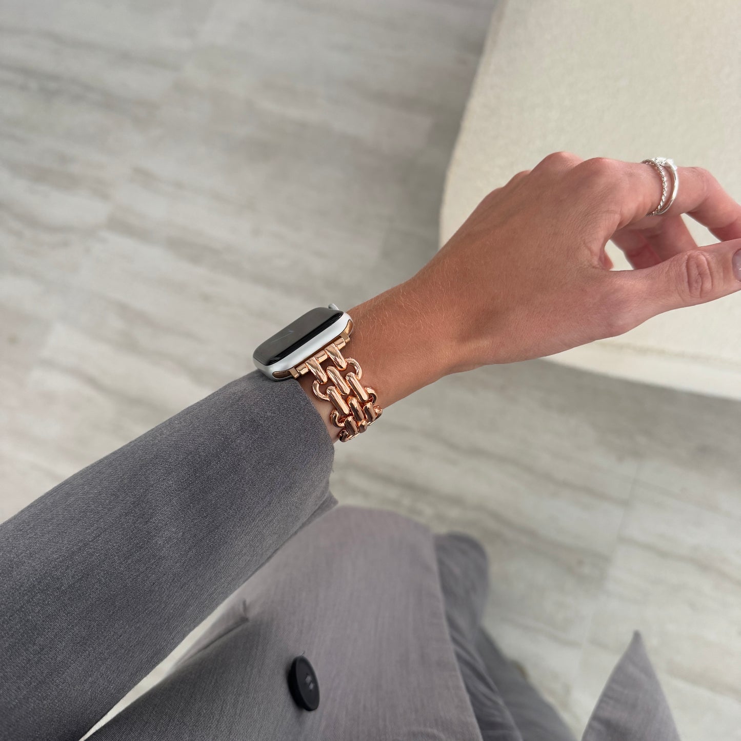 Rose gold chain link Apple Watch band worn on a wrist above grey trousers. Fashionable metal iWatch strap, compatible with Apple Watch 38mm to 49mm, Series 1-11, SE and Ultra