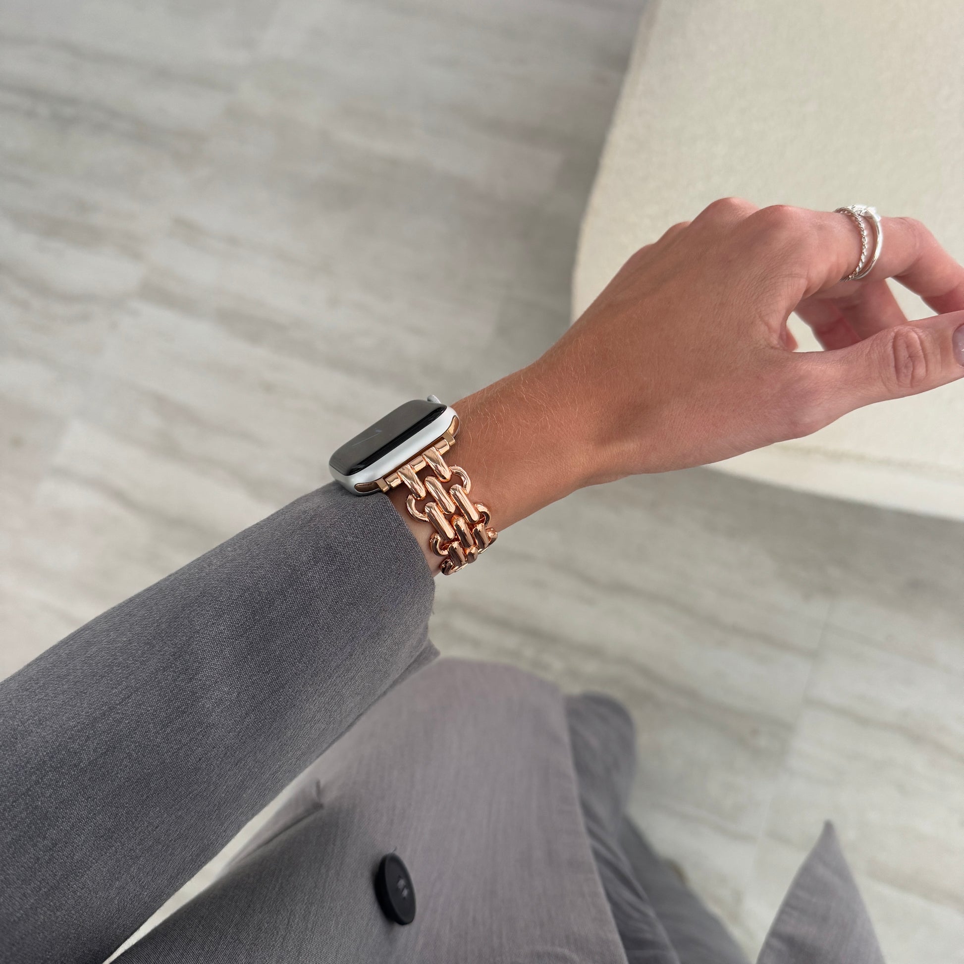 Rose gold chain link Apple Watch band worn on a wrist above grey trousers. Fashionable metal iWatch strap, compatible with Apple Watch 38mm to 49mm, Series 1-11, SE and Ultra