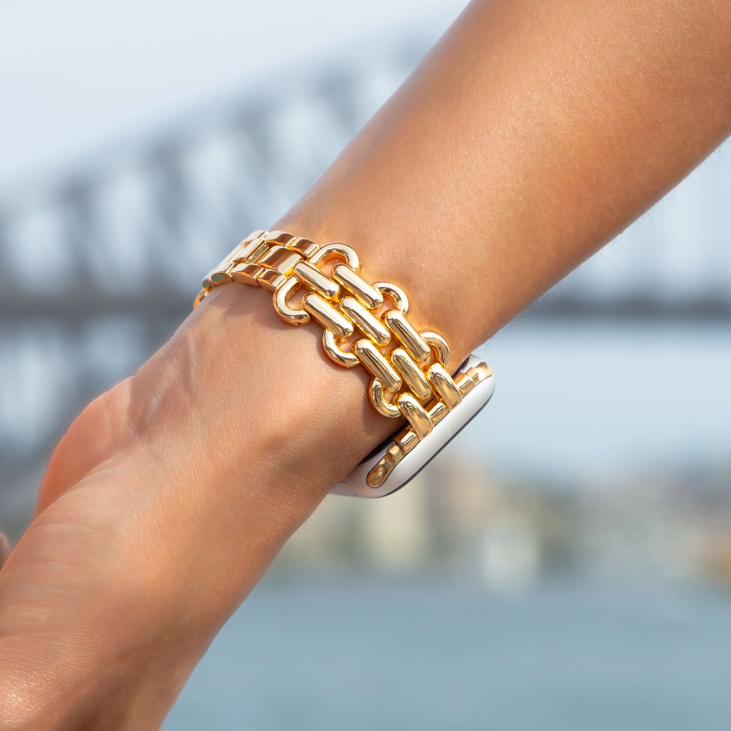 Gold chain link Apple Watch band worn outdoors by a harbour bridge. Sleek metal iWatch strap, compatible with Apple Watch 38mm to 49mm, Series 1-11, SE and Ultra