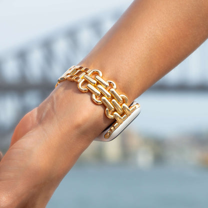 Gold chain link Apple Watch band worn outdoors by a harbour bridge. Sleek metal iWatch strap, compatible with Apple Watch 38mm to 49mm, Series 1-11, SE and Ultra