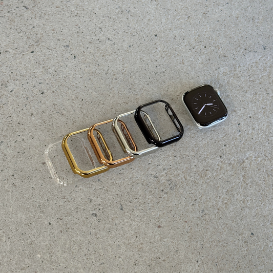 Five smartwatch bezel cases in gold, rose gold, silver, and black are arranged in a row on a textured concrete surface, with one Apple watch shown next to the black case.