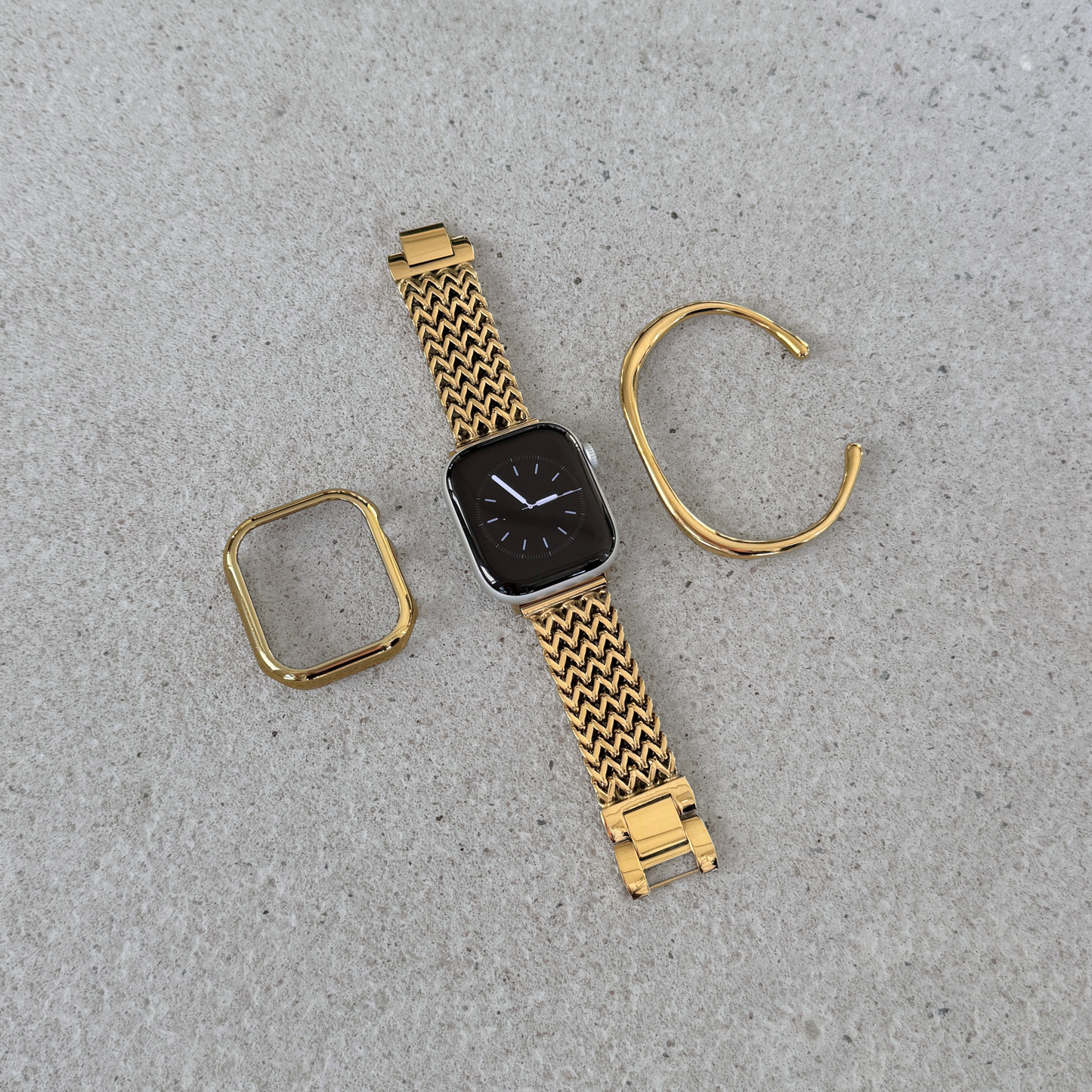 Gold tone metal Apple watch band, matching watch case and cuff bracelet. Components arranged on a light textured surface. The band has a woven zigzag pattern and includes a watch face with a dark display.