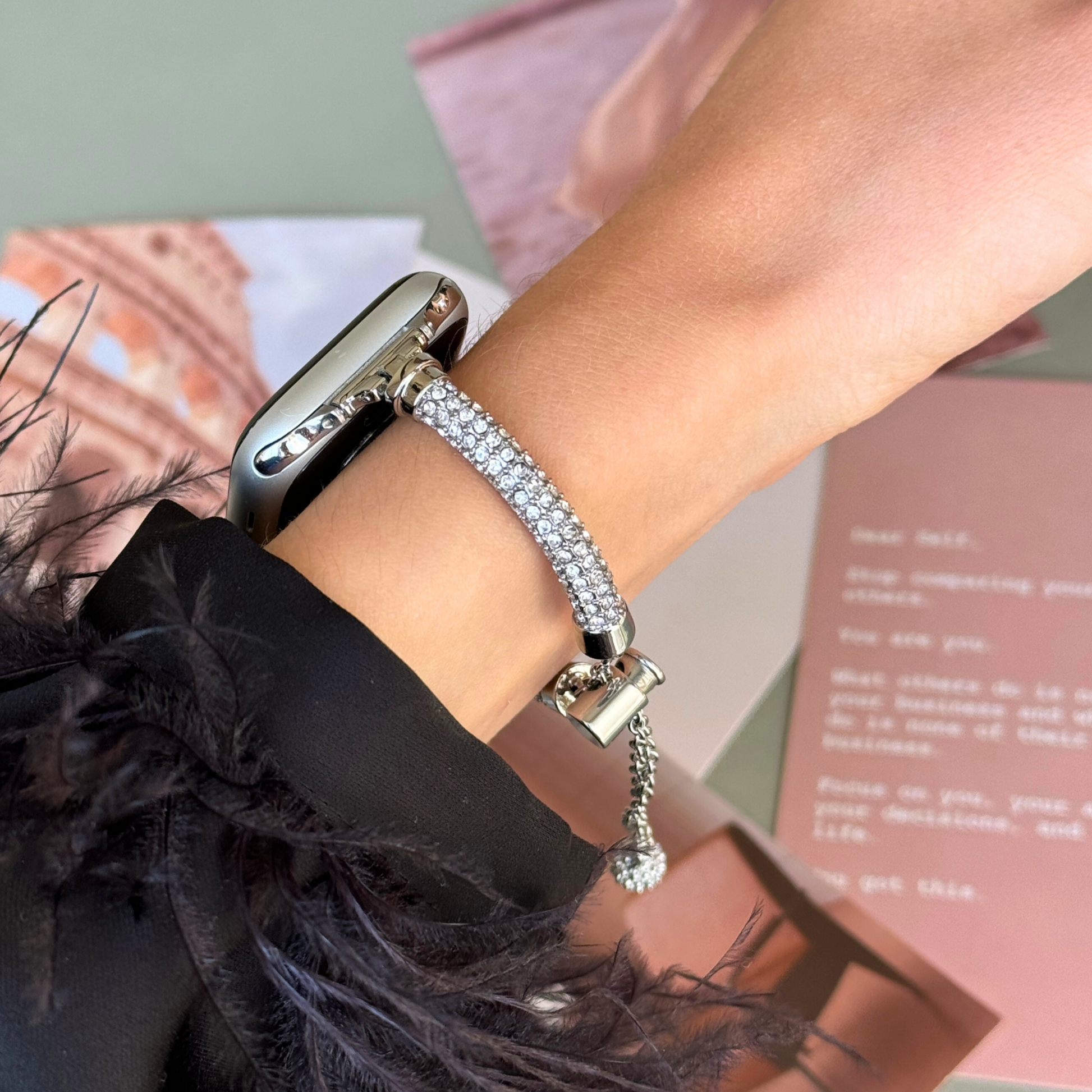 Silver rhinestone Apple Watch band worn on a wrist above pink and green papers, metal strap for Apple Watch 38mm to 49mm