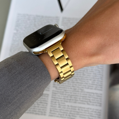 Gold slim link Apple Watch band worn with a grey sleeve over a newspaper, stainless steel bracelet for Apple Watch 38mm to 49mm