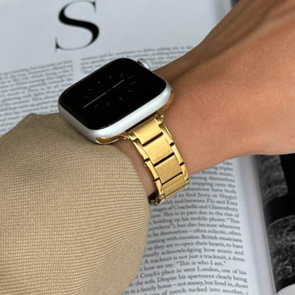 Gold slim link Apple Watch band worn with a camel sleeve above an open book, stainless steel bracelet for Apple Watch 38mm to 49mm