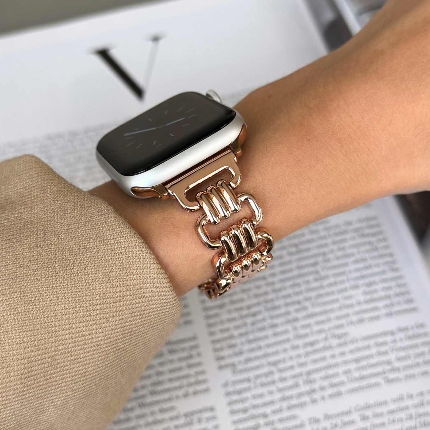 Rose gold open link Apple Watch band worn with a camel sleeve above an open book. Elegant metal iWatch armband, compatible with Apple Watch 38mm to 49mm, Series 1-11, SE and Ultra