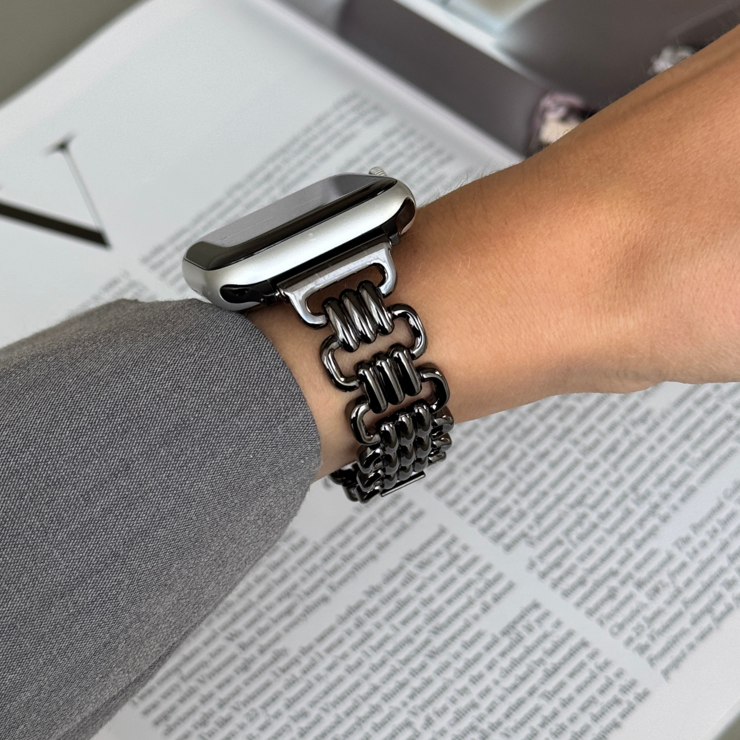 Black open link Apple Watch band worn with a grey sleeve over a newspaper. High-end metal iWatch strap, compatible with Apple Watch 38mm to 49mm, Series 1-11, SE and Ultra