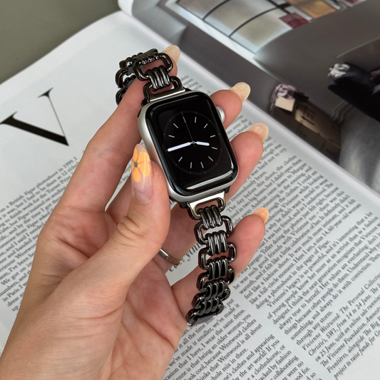 Black open link Apple Watch band held in a hand over a magazine page. Elegant metal iWatch bracelet, compatible with Apple Watch 38mm to 49mm, Series 1-11, SE and Ultra