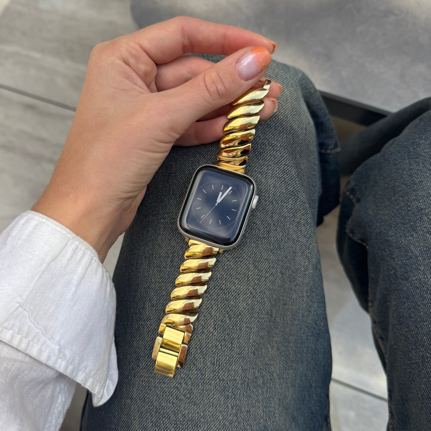 Gold twisted link Apple Watch band worn on a wrist resting on denim. Feminine stainless steel iWatch strap with a dynamic silhouette, compatible with Apple Watch 38mm to 49mm, Series 1-11, SE and Ultra