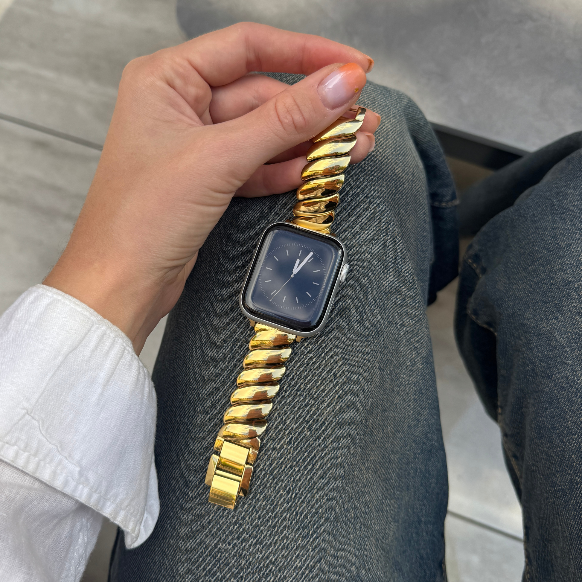 Gold twisted link Apple Watch band worn on a wrist resting on denim. Feminine stainless steel iWatch strap with a dynamic silhouette, compatible with Apple Watch 38mm to 49mm, Series 1-11, SE and Ultra