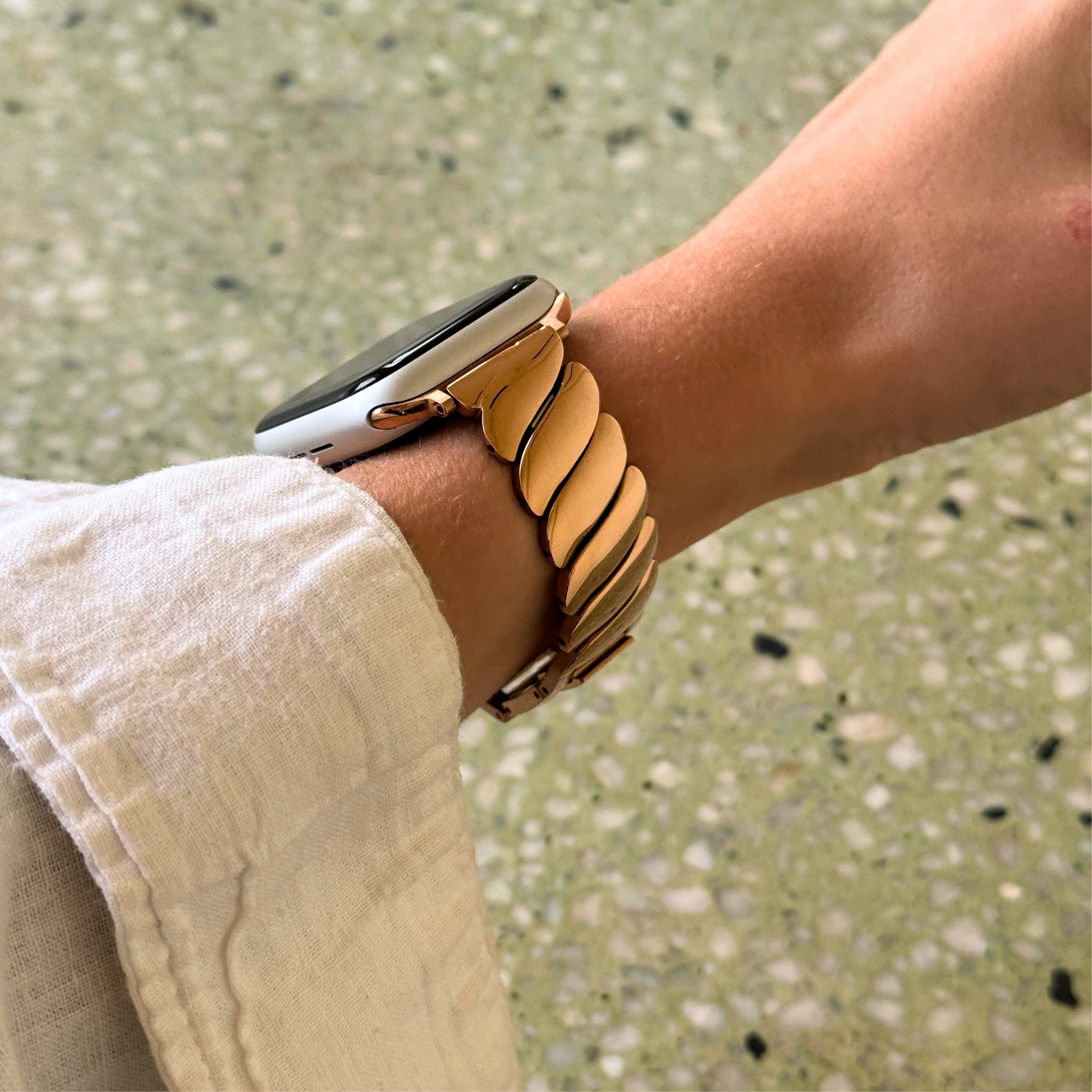 Rose gold twisted link Apple Watch band worn on a wrist with a cream linen sleeve. Bold refined stainless steel iWatch bracelet, compatible with Apple Watch 38mm to 49mm, Series 1-11, SE and Ultra