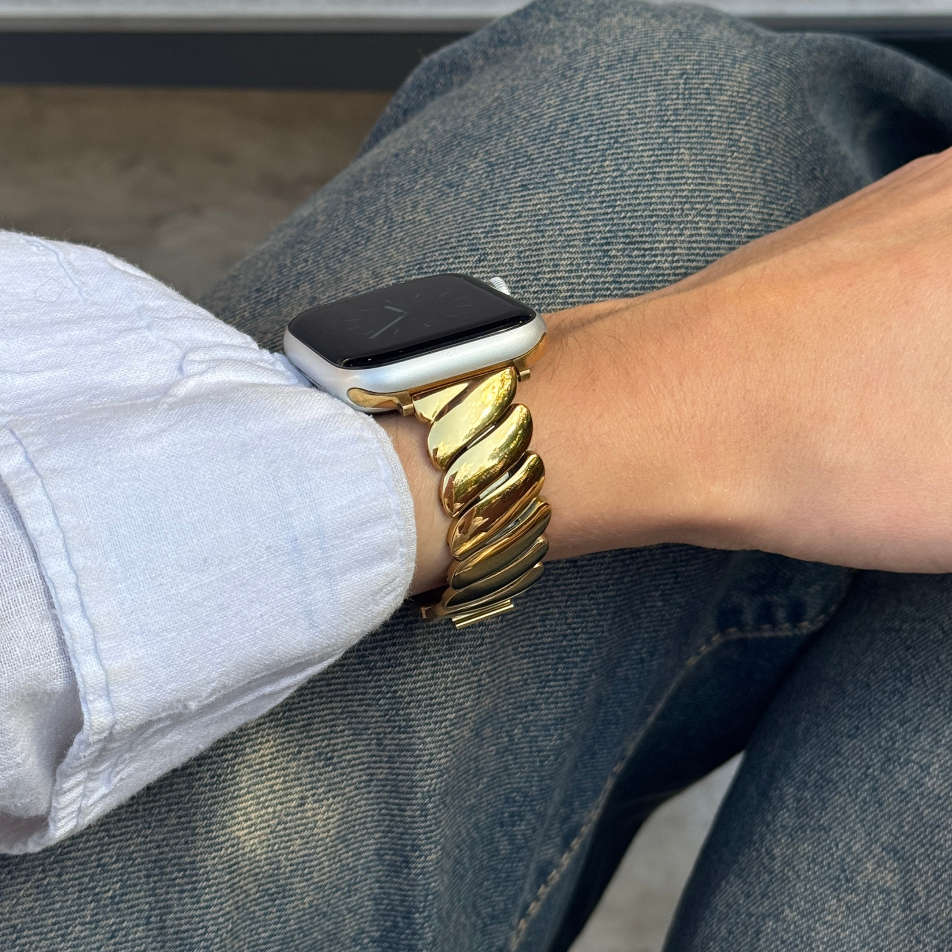 Gold twisted link Apple Watch band worn with a white shirt sleeve over denim. Premium stainless steel iWatch armband, compatible with Apple Watch 38mm to 49mm, Series 1-11, SE and Ultra