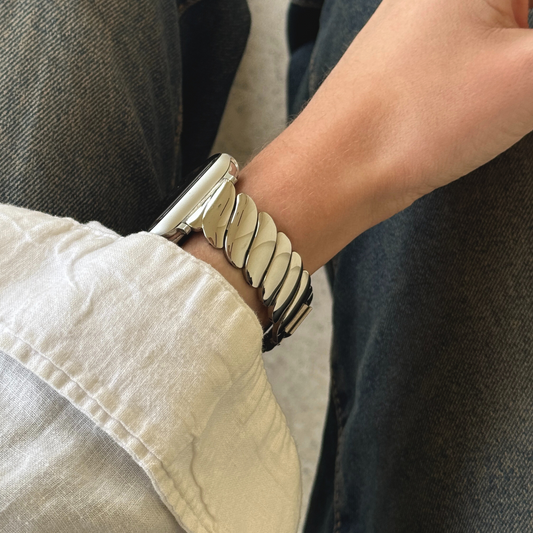 Silver twisted link Apple Watch band worn with a pale sleeve over denim. Feminine stainless steel iWatch strap with a unique twisted design, compatible with Apple Watch 38mm to 49mm, Series 1-11, SE and Ultra