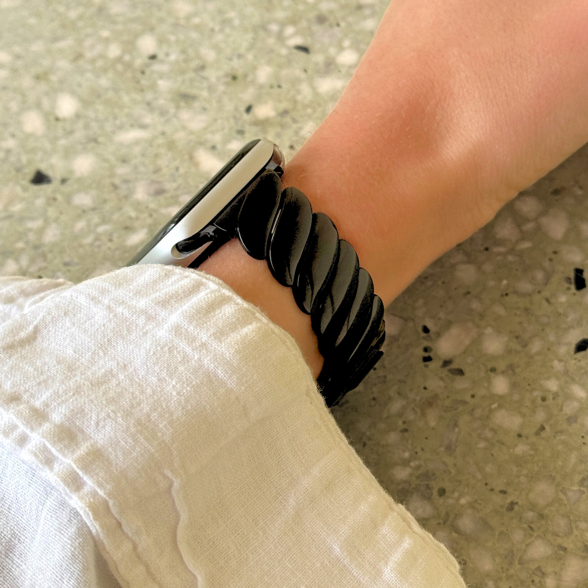 Black twisted link Apple Watch band worn on a wrist with a white linen sleeve. Waterproof stainless steel iWatch bracelet, compatible with Apple Watch 38mm to 49mm, Series 1-11, SE and Ultra