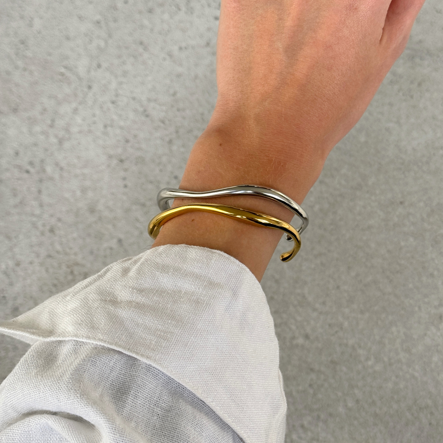 Two thin wavy cuff bracelets, one silver and one gold, worn on a person's wrist with a linen sleeve visible.