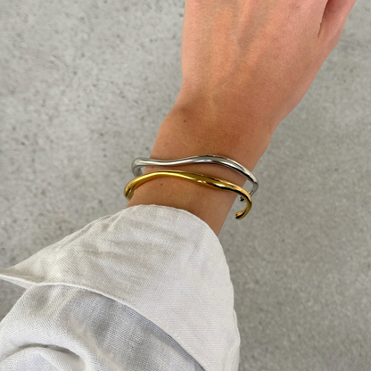 Two thin wavy cuff bracelets, one silver and one gold, worn on a person's wrist with a linen sleeve visible.