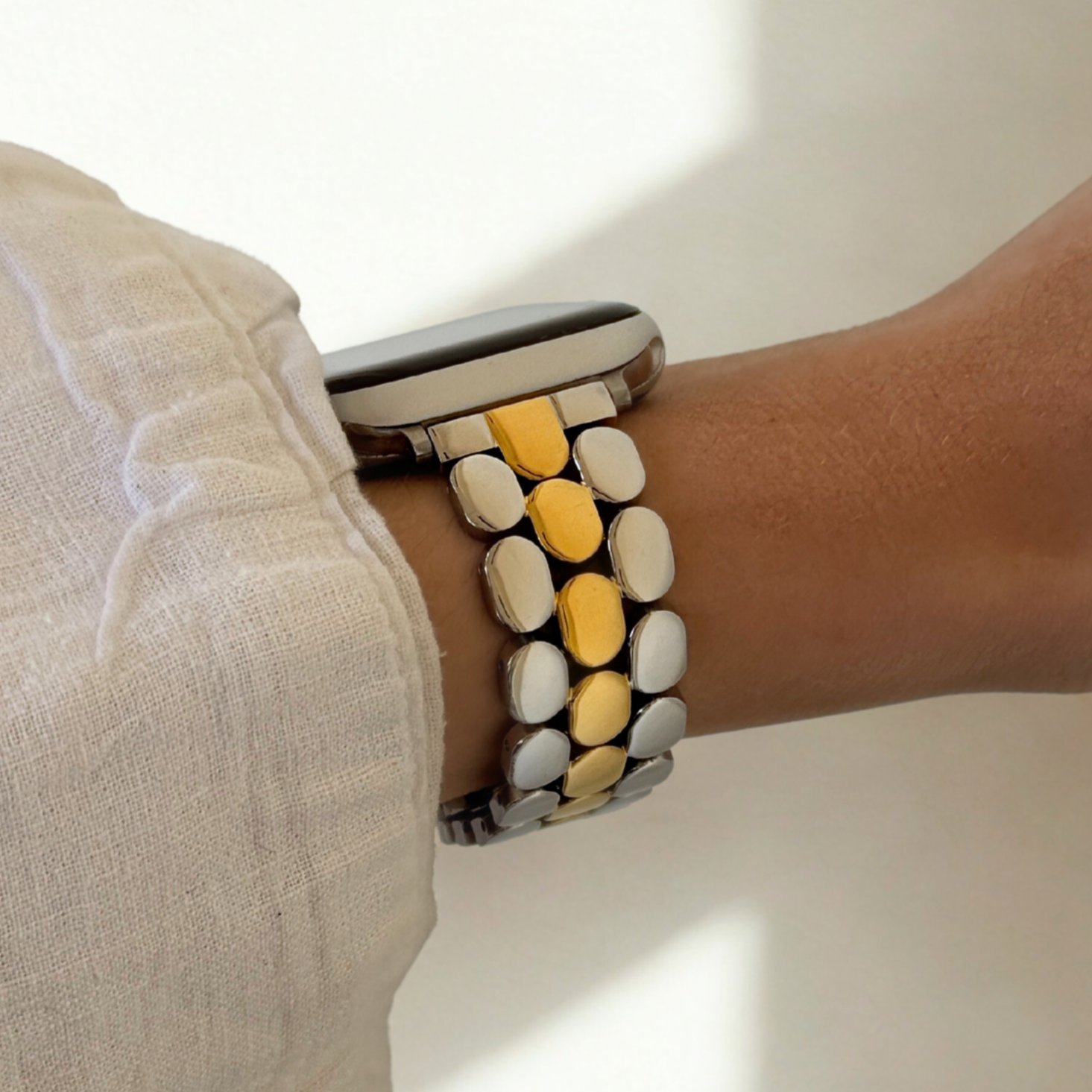 Silver gold pebble link Apple Watch band worn on a wrist with a white linen cuff. Slim high shine stainless steel iWatch bracelet with a butterfly clasp, compatible with Apple Watch 38mm, 40mm, 41mm, 42mm, 44mm, 45mm, 46mm and 49mm, Series 1-11, SE and Ultra