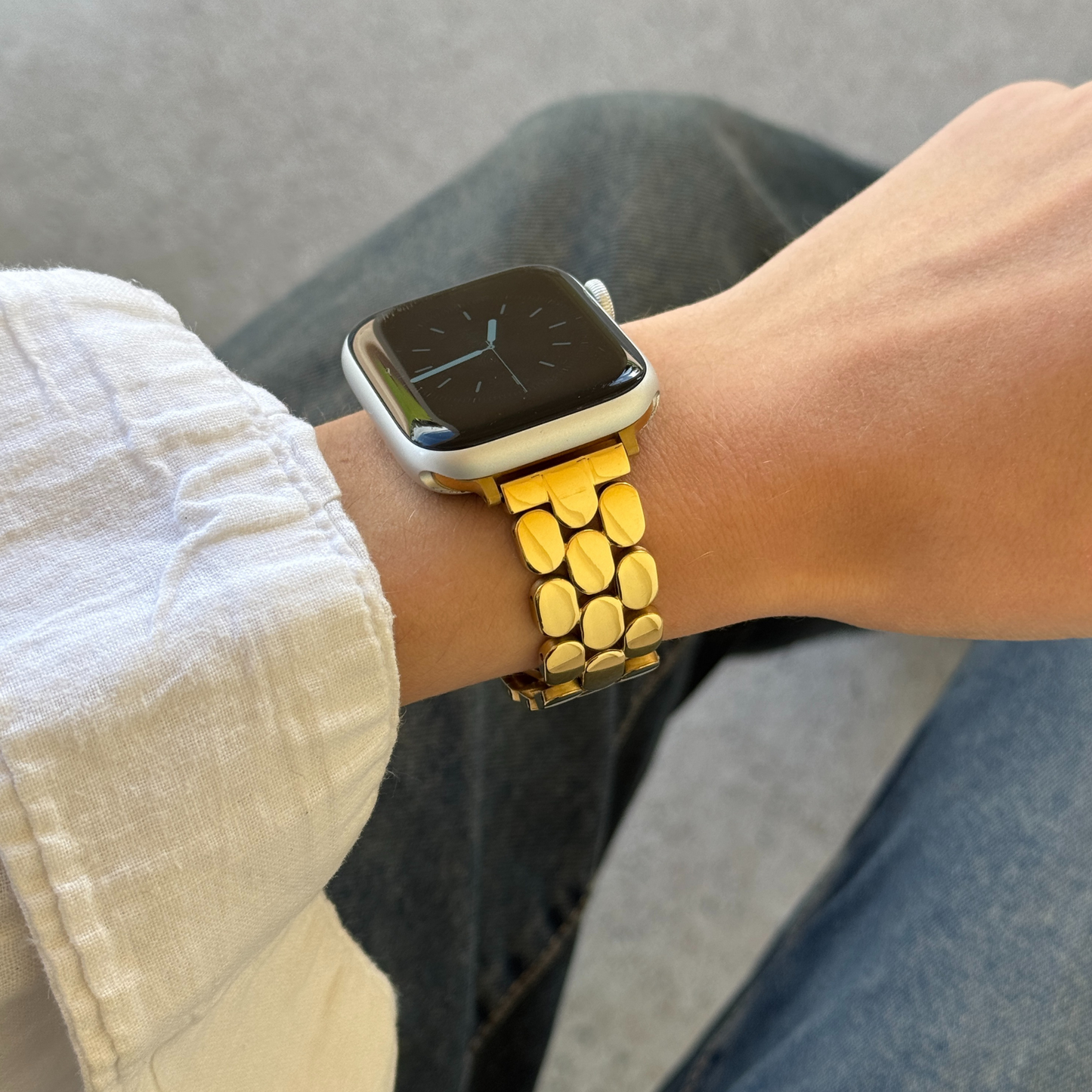 Gold pebble link Apple Watch band worn on a wrist above grey trousers. Feminine stainless steel iWatch bracelet with a high shine finish, compatible with Apple Watch 38mm to 49mm, Series 1-11, SE and Ultra
