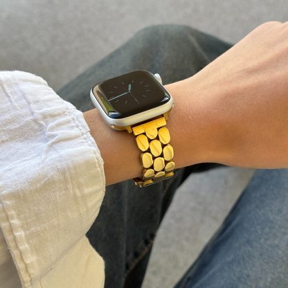 Gold pebble link Apple Watch band worn on a wrist above grey trousers. Feminine stainless steel iWatch bracelet with a high shine finish, compatible with Apple Watch 38mm to 49mm, Series 1-11, SE and Ultra