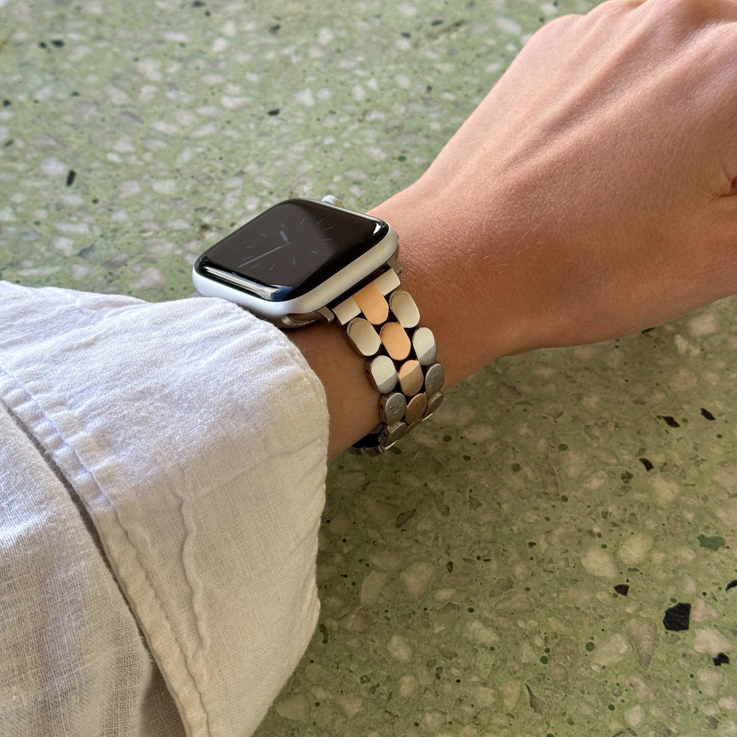 Silver rose gold pebble link Apple Watch band worn on a wrist against a green terrazzo background. Modern stainless steel iWatch armband, compatible with Apple Watch 38mm to 49mm, Series 1-11, SE and Ultra