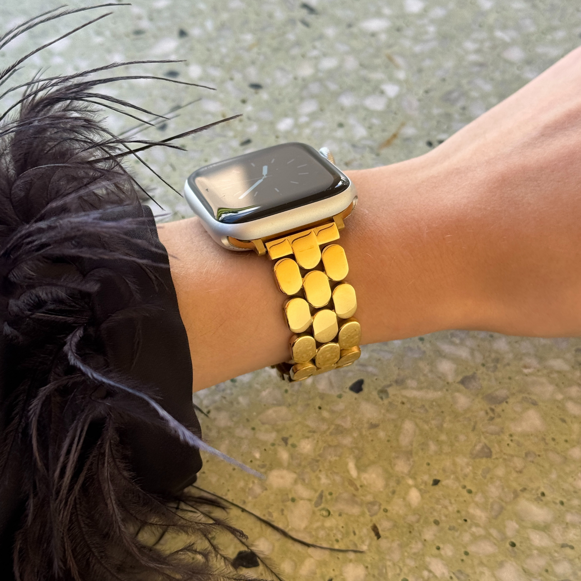 Gold pebble link Apple Watch band worn with a feather trimmed cuff outdoors. Timeless stainless steel iWatch strap with a high shine finish, compatible with Apple Watch 38mm to 49mm, Series 1-11, SE and Ultra