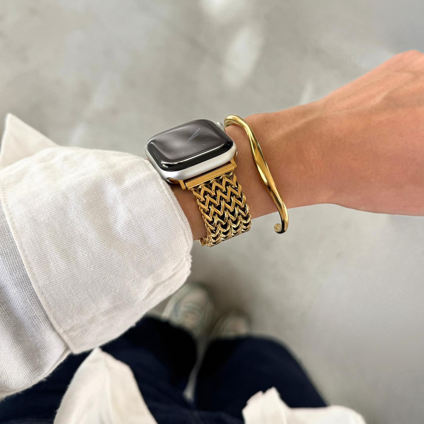 A close-up of a person's wrist wearing a gold toned mesh Apple watch band, along with a gold bangle bracelet and a linen shirt.