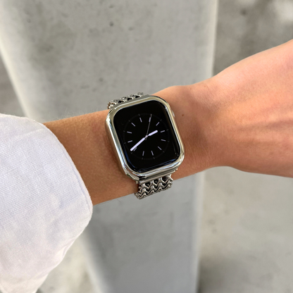A close-up of a person's wrist wearing a silver toned metal link Apple watch with a rectangular black screen and a polished silver bezel, set against a blurred  concrete background.