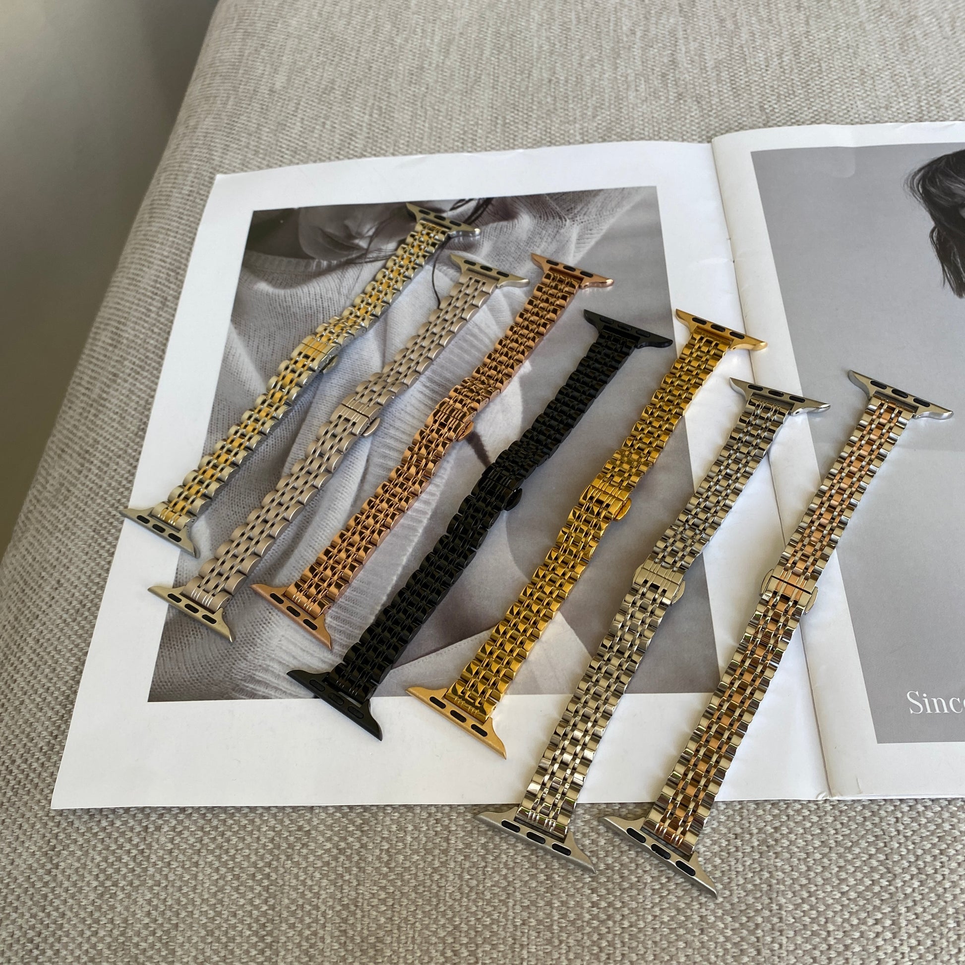 Silver, gold, rose gold, black, starlight, silver gold and silver rose gold woven link Apple Watch bands laid out on a magazine, stainless steel straps for Apple Watch 38mm to 49mm
