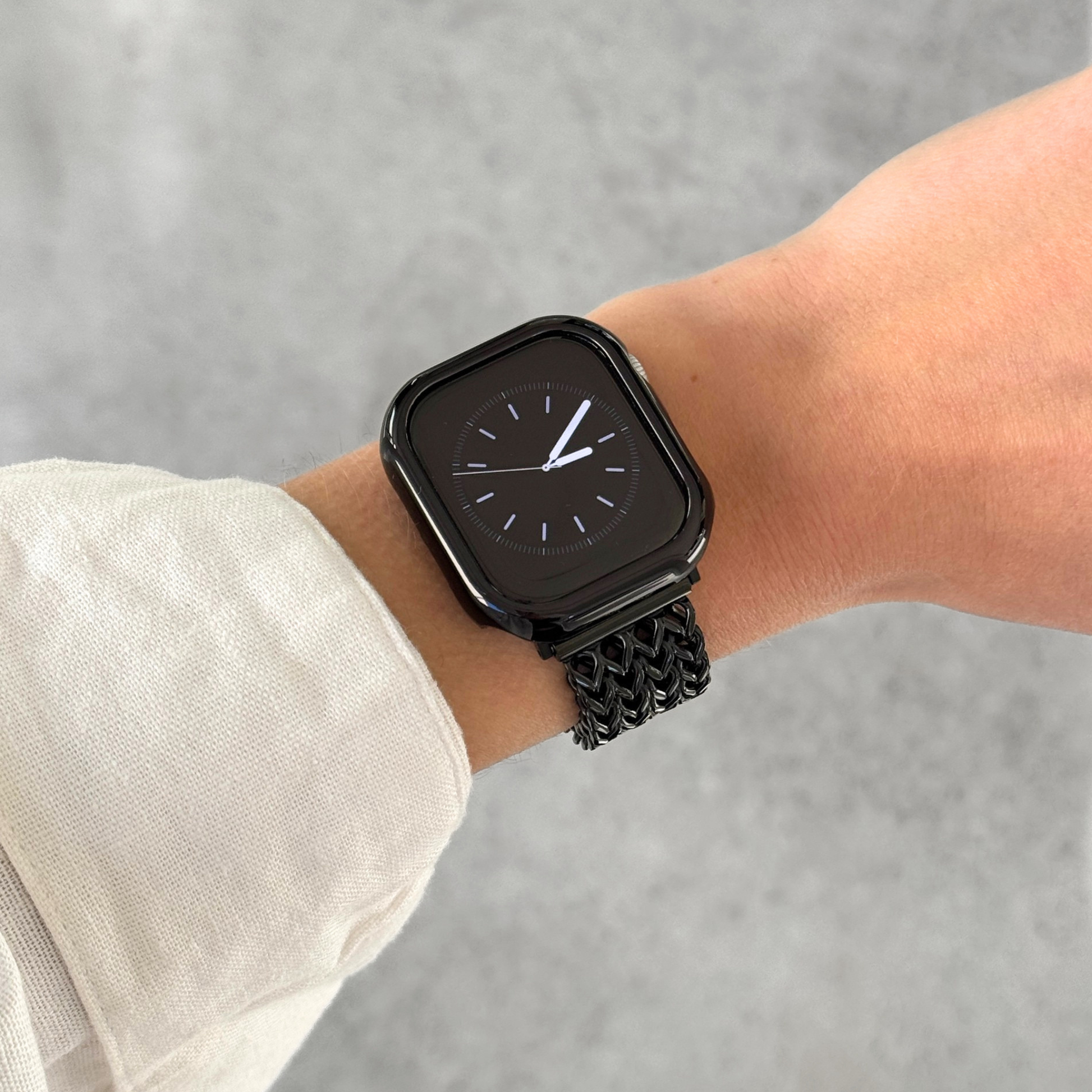 A close-up of a person's wrist wearing a black Apple smartwatch with a black mesh band and a black metallic bumper case cover, against a light textured background.