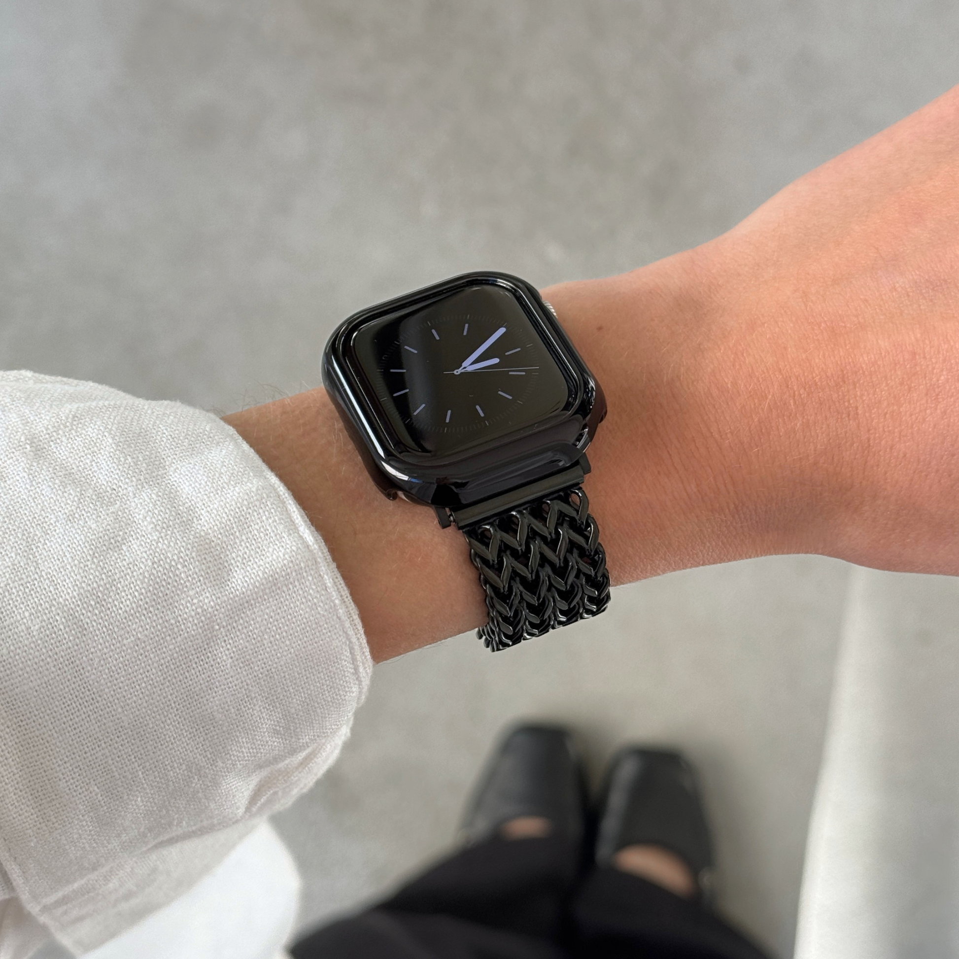 A close-up of a person's wrist wearing a black Apple smartwatch with a black mesh band and a matching bezel, set against a concrete background with shoes visible at the bottom.
