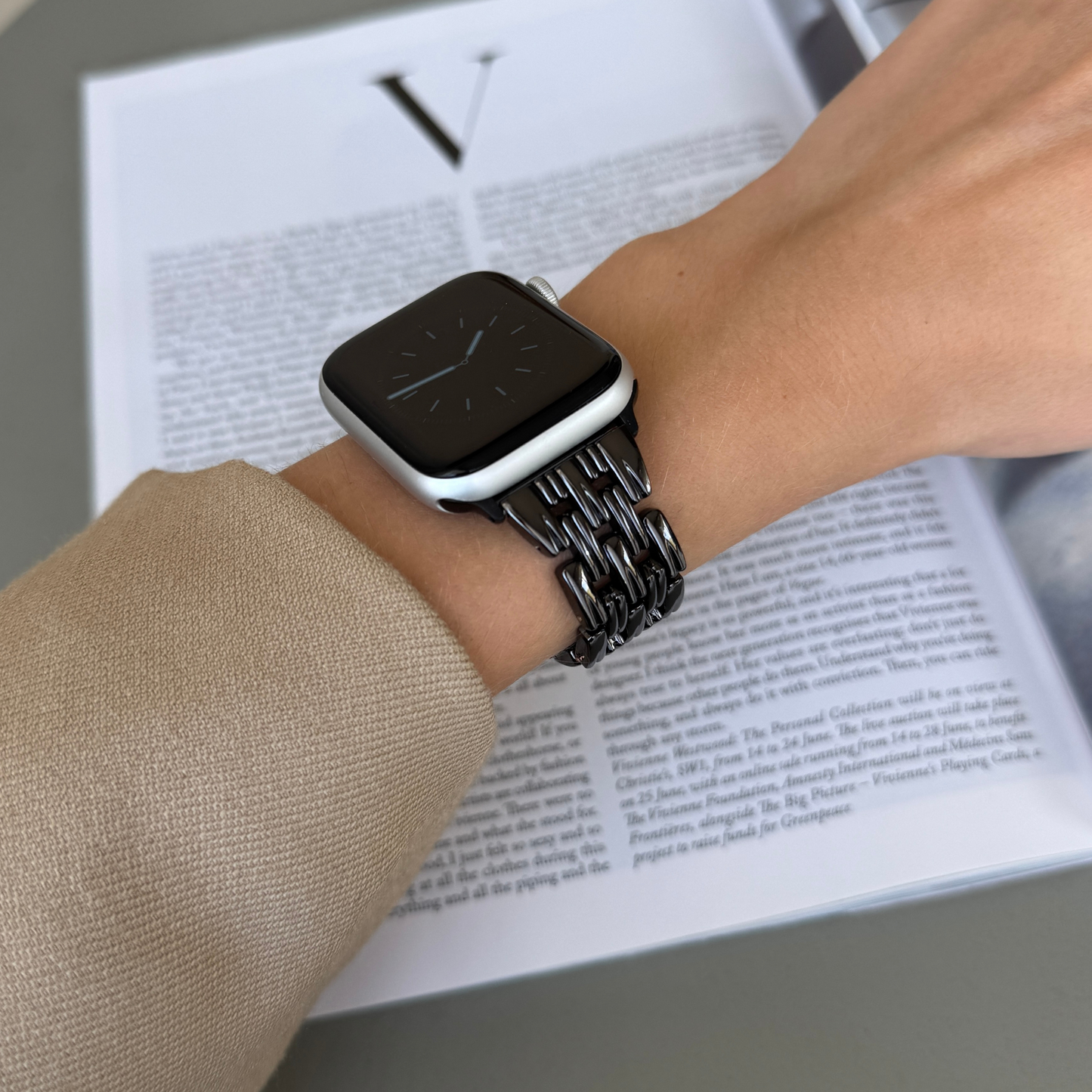Black link Apple Watch band worn with a beige sleeve above an open magazine. Metal iWatch strap for women, compatible with Apple Watch 38mm to 49mm, Series 1-11, SE and Ultra