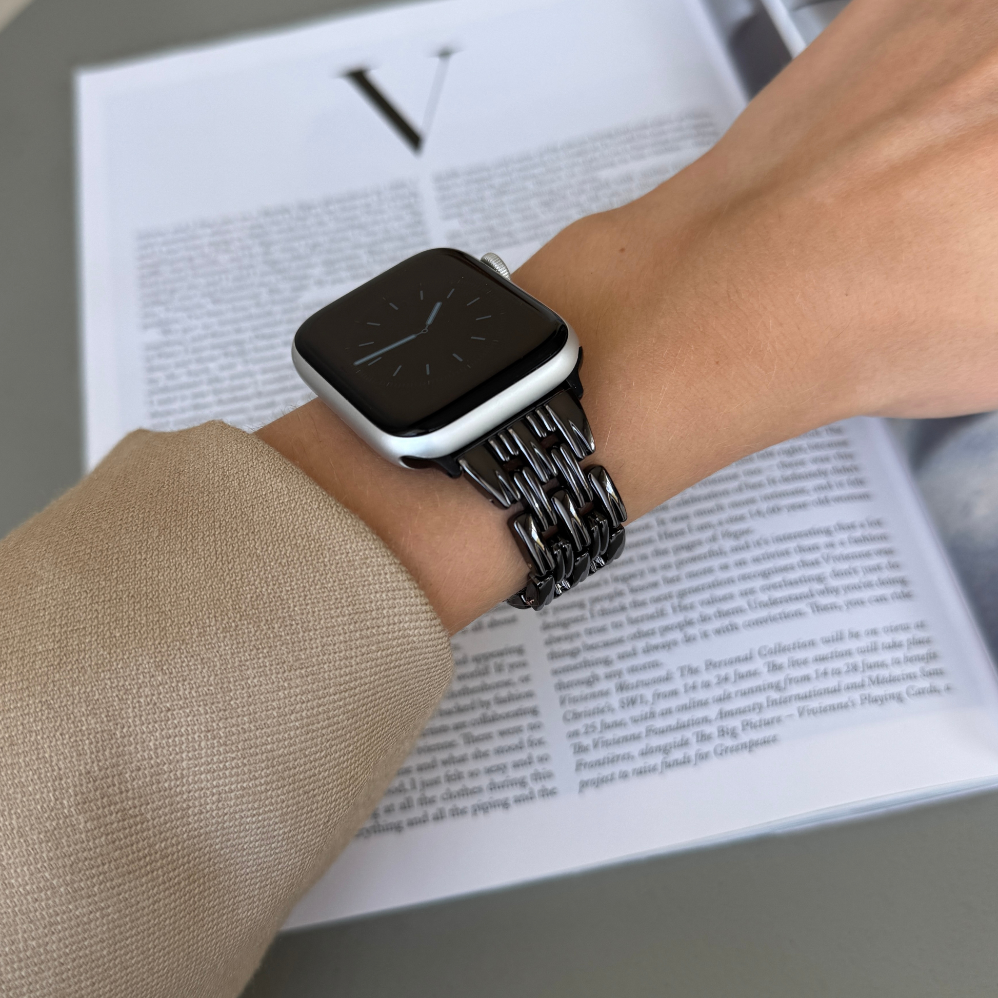 Black link Apple Watch band worn with a beige sleeve above an open magazine. Metal iWatch strap for women, compatible with Apple Watch 38mm to 49mm, Series 1-11, SE and Ultra
